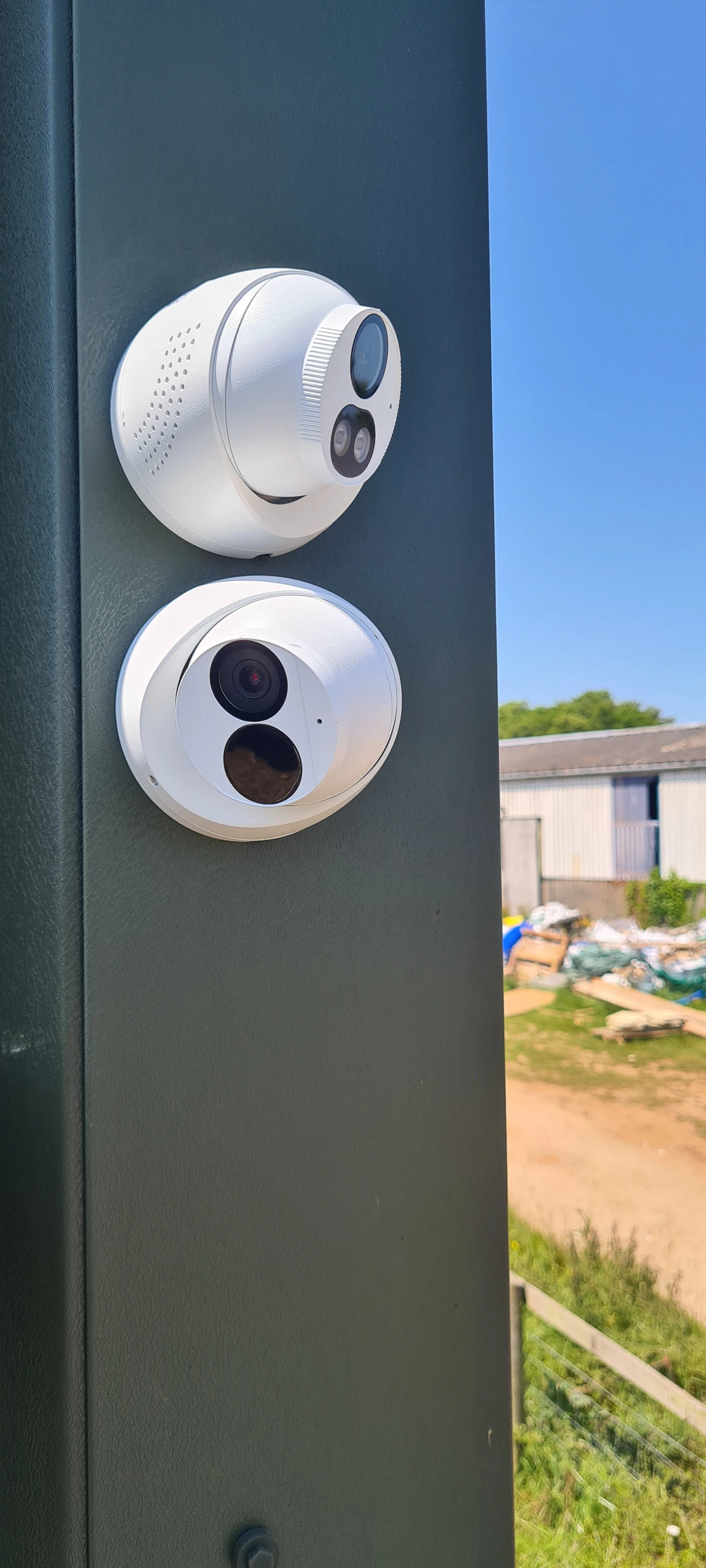 Two security cameras mounted on a green pole, with a clear blue sky in the background and a blurred structure visible below.