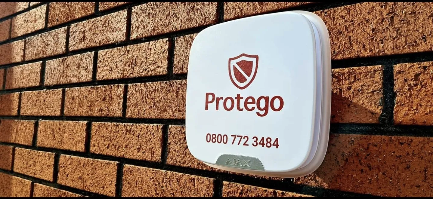 A white security device mounted on a textured brown brick wall, displaying "Protego" and a phone number.