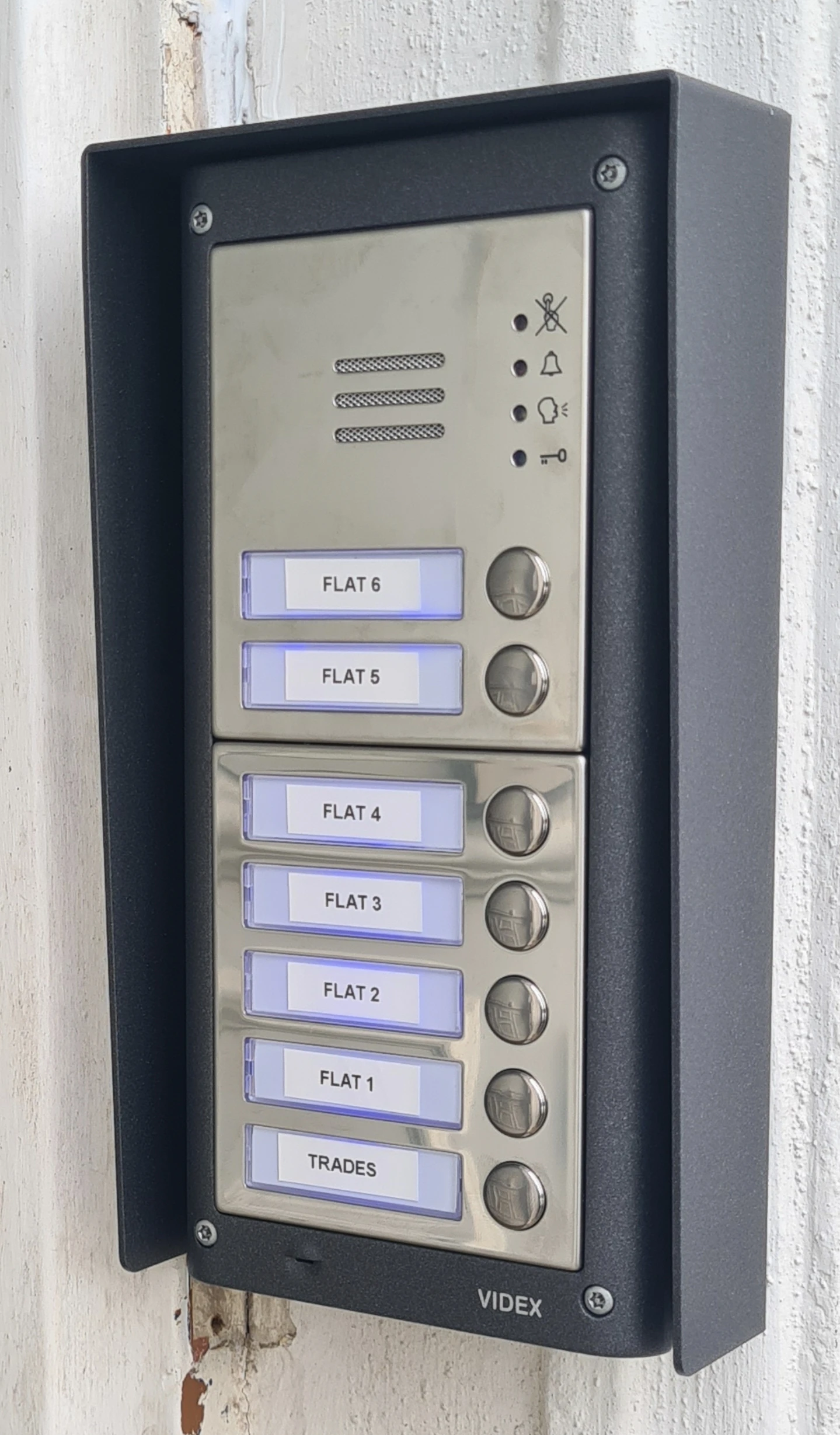 Metal intercom panel with buttons for eight flats and trades, labeled and lit, mounted on a wall.