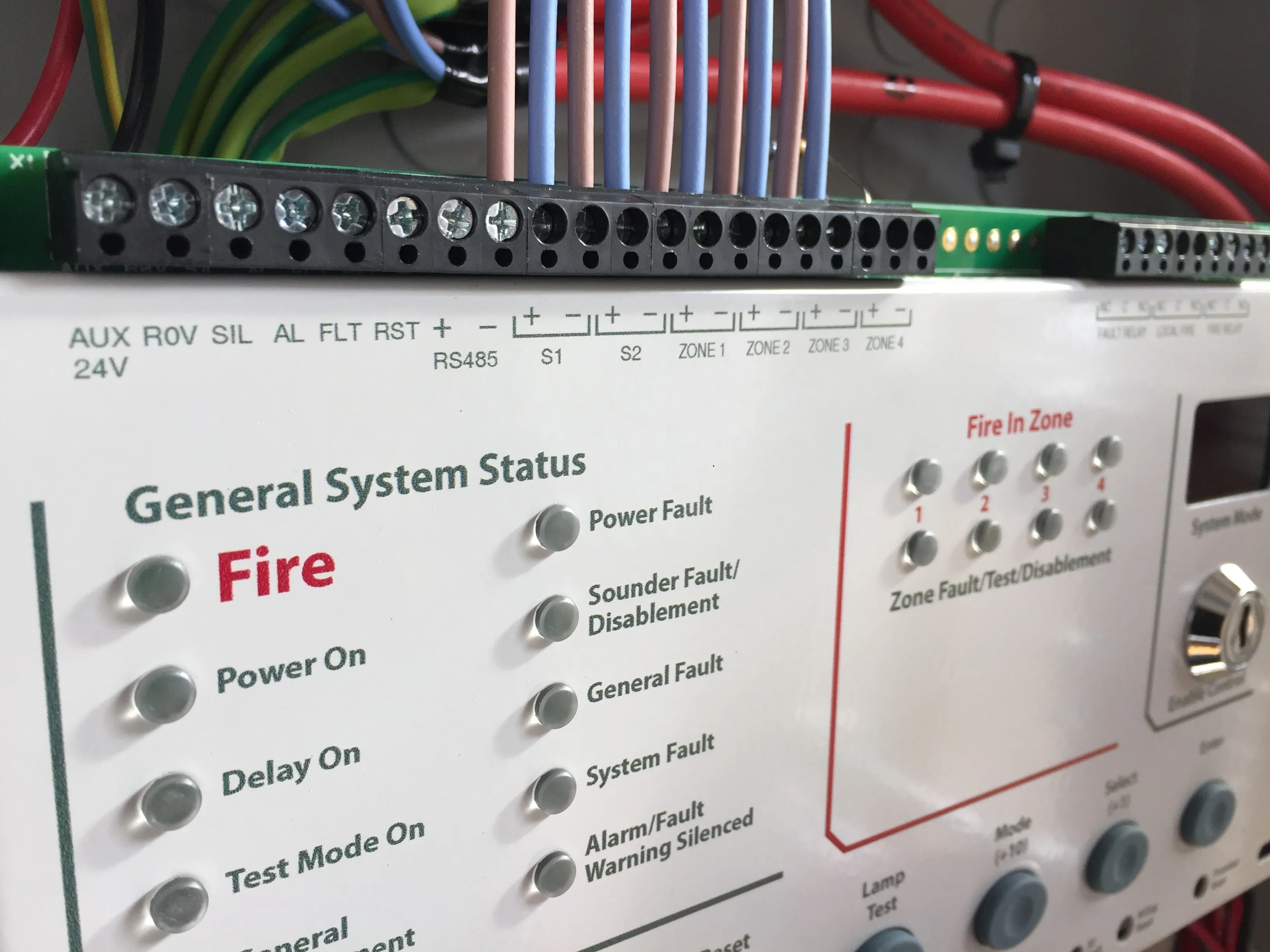 Control panel of a fire alarm system showing indicators for fire status, power, and zone tests with wiring connections above.