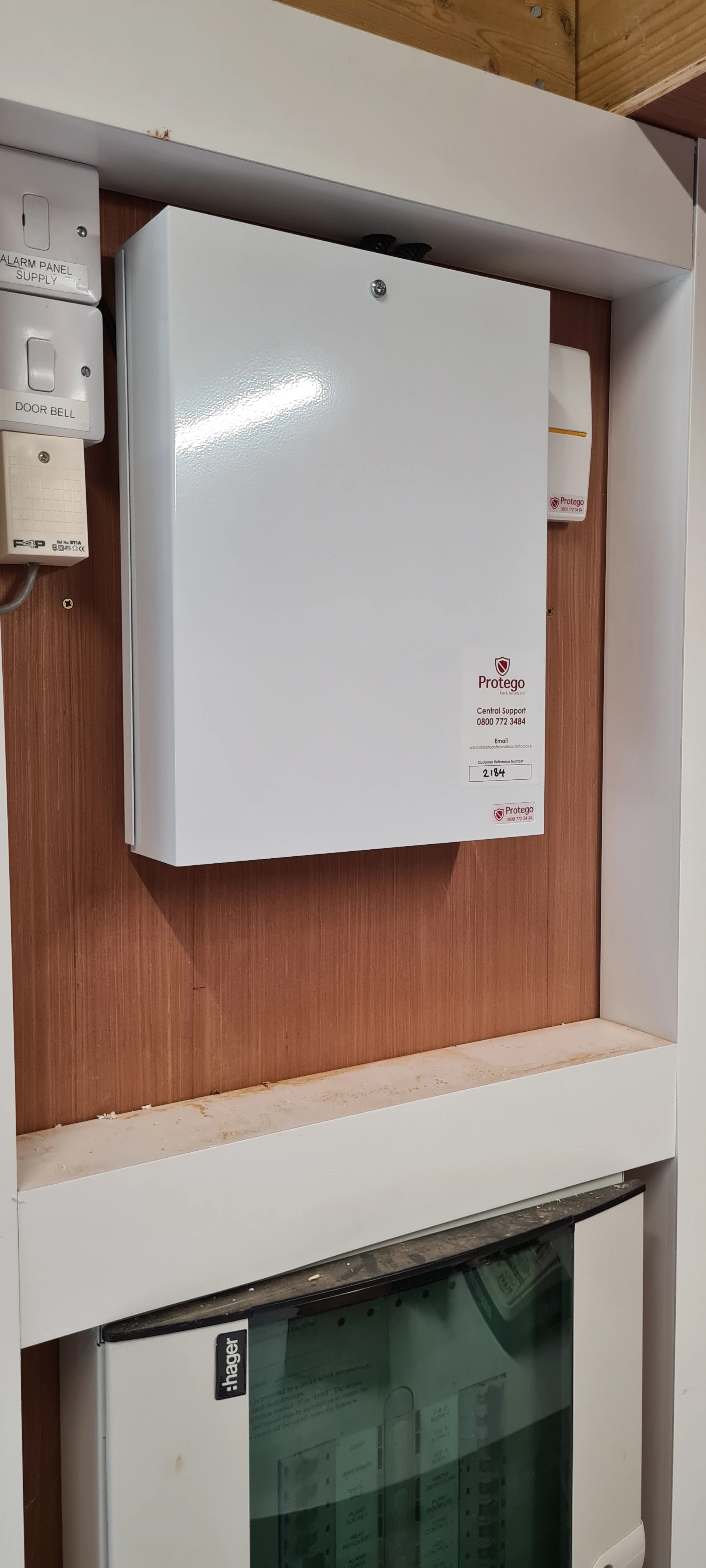 White electrical cabinet mounted on a wooden wall, with labels for alarm and doorbell controls above.