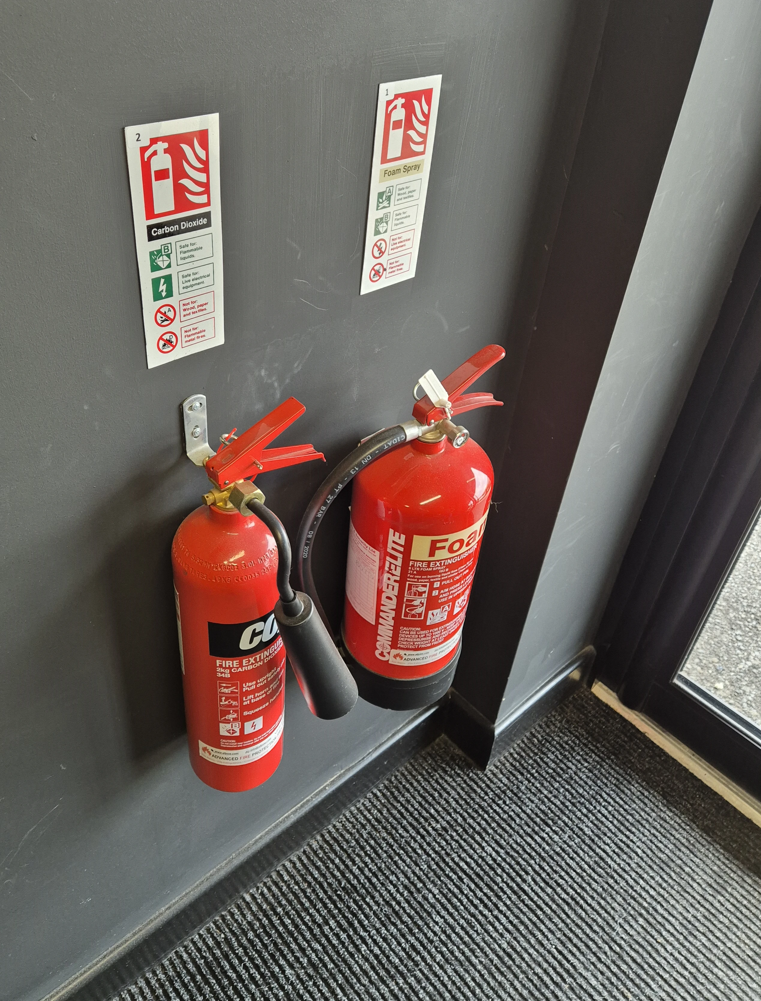 Two fire extinguishers mounted on a wall: one labeled "Foam Spray" and the other "Carbon Dioxide," with safety signage nearby.