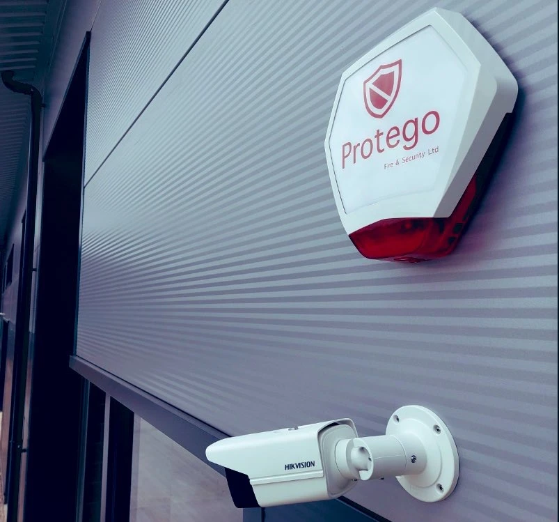 Security camera mounted on a wall next to a fire alarm sign from Protego Fire & Security Ltd. Modern building exterior.