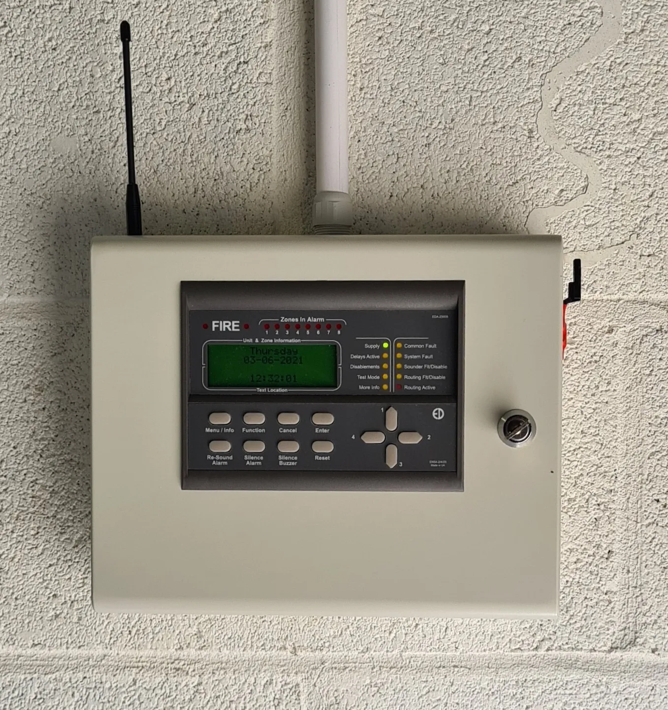 A fire alarm control panel mounted on a wall, displaying the date and various indicator lights and control buttons.