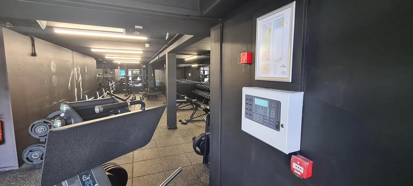 View into a gym with weights and equipment. A fire alarm panel and a layout map are mounted on the wall.
