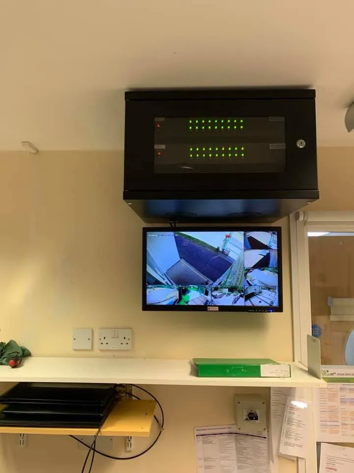 Monitor displaying multiple camera feeds mounted above a black device with green indicator lights. Shelves with equipment below.