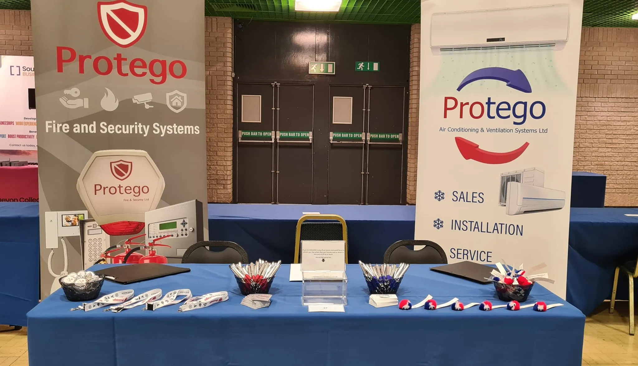 Exhibition booth for Protego featuring banners for fire, security, and HVAC services, with promotional items on a table.