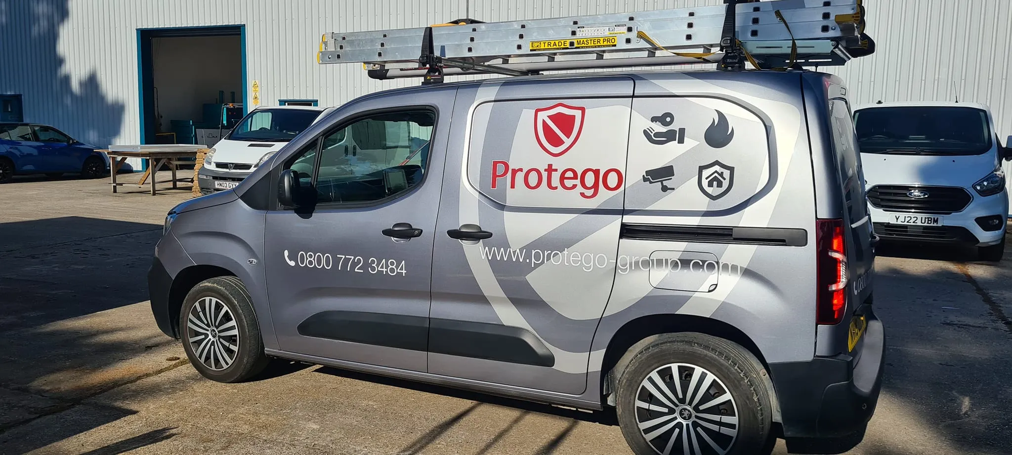 Silver service van with "Protego" branding, safety icons, and a ladder on top, parked in a lot with other vehicles and a building in background.
