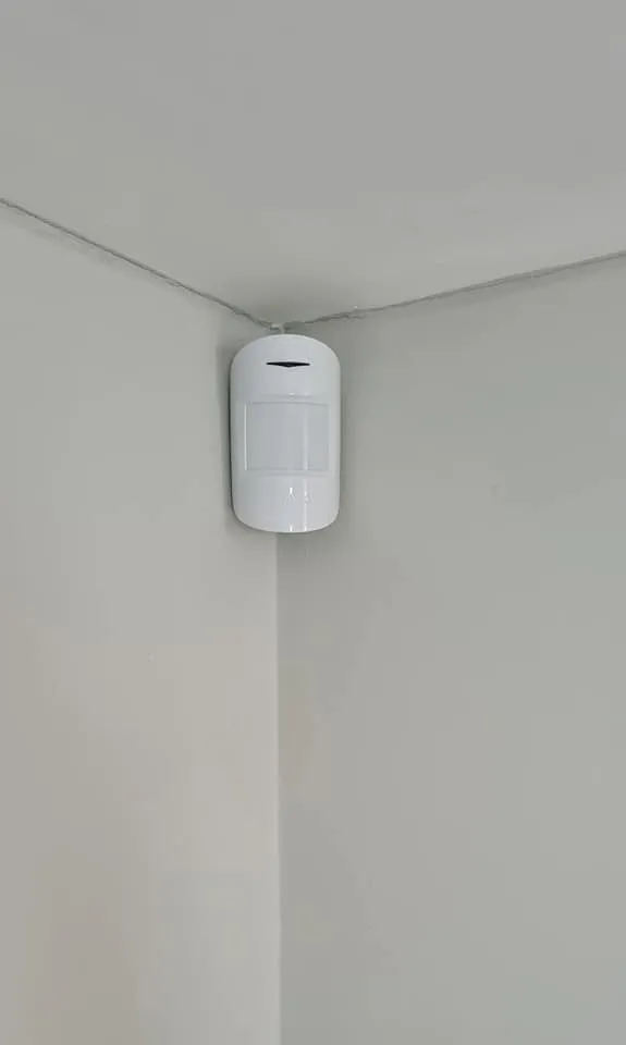 White motion sensor mounted in a corner on a light-colored wall, designed for security or automation purposes.