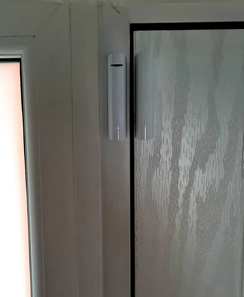 Close-up of a white door frame with a door sensor mounted on the left side, adjacent to a glass door with textured surface.