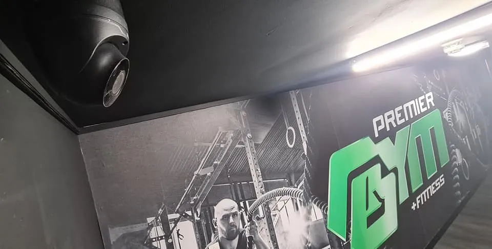 Interior of a gym with a wall mural featuring fitness equipment and the "Premier Gym" logo in green, along with a mounted speaker.