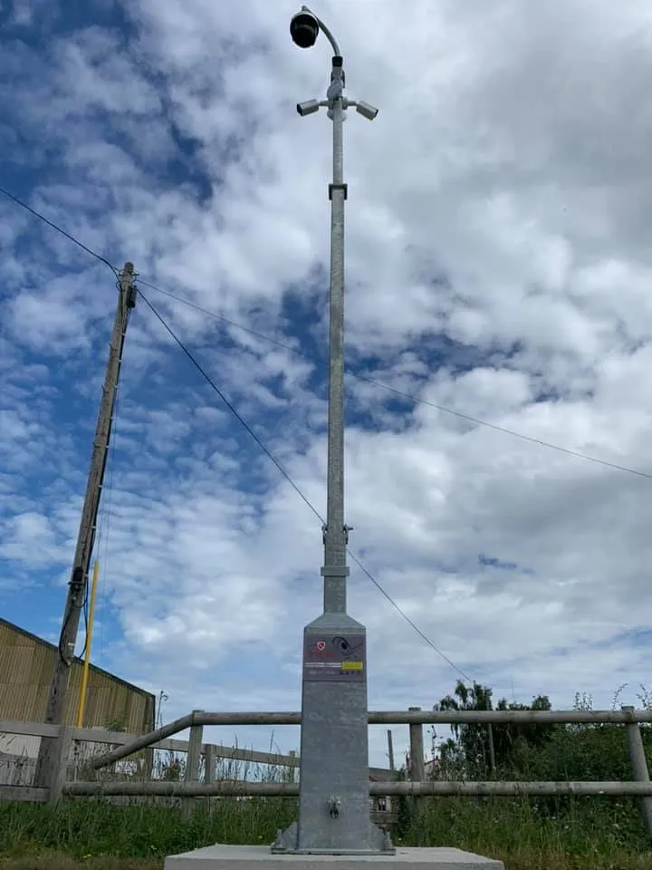 Metal pole with surveillance camera mounted on top, set against a cloudy blue sky and grassy area.
