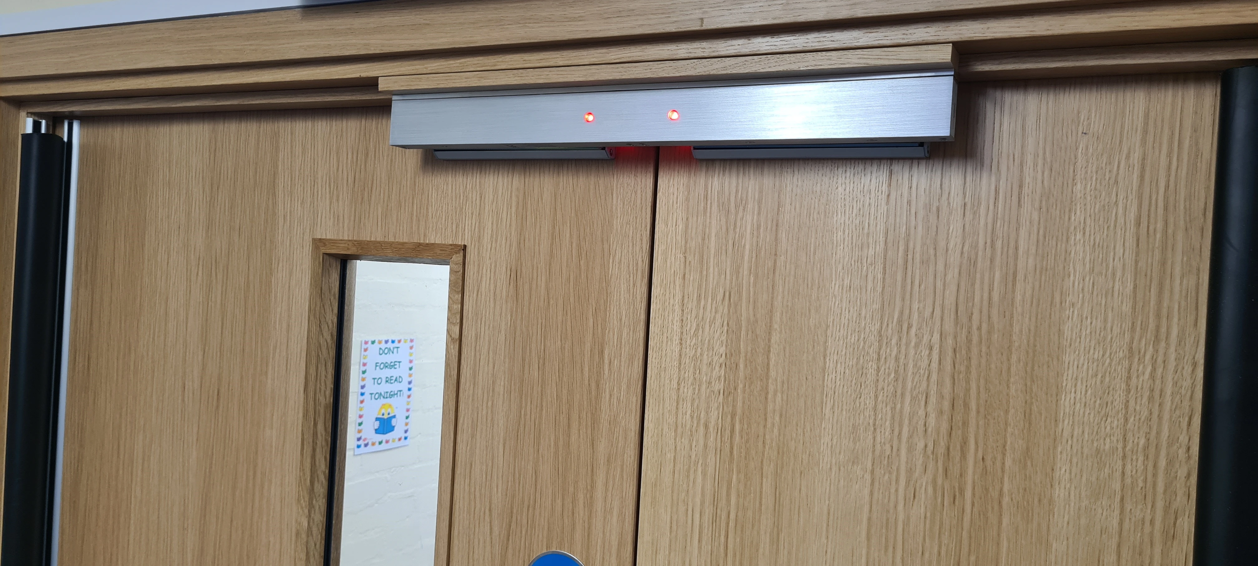 Automatic door system with red indicator lights above wooden doors, next to a small window and a colorful reminder note.